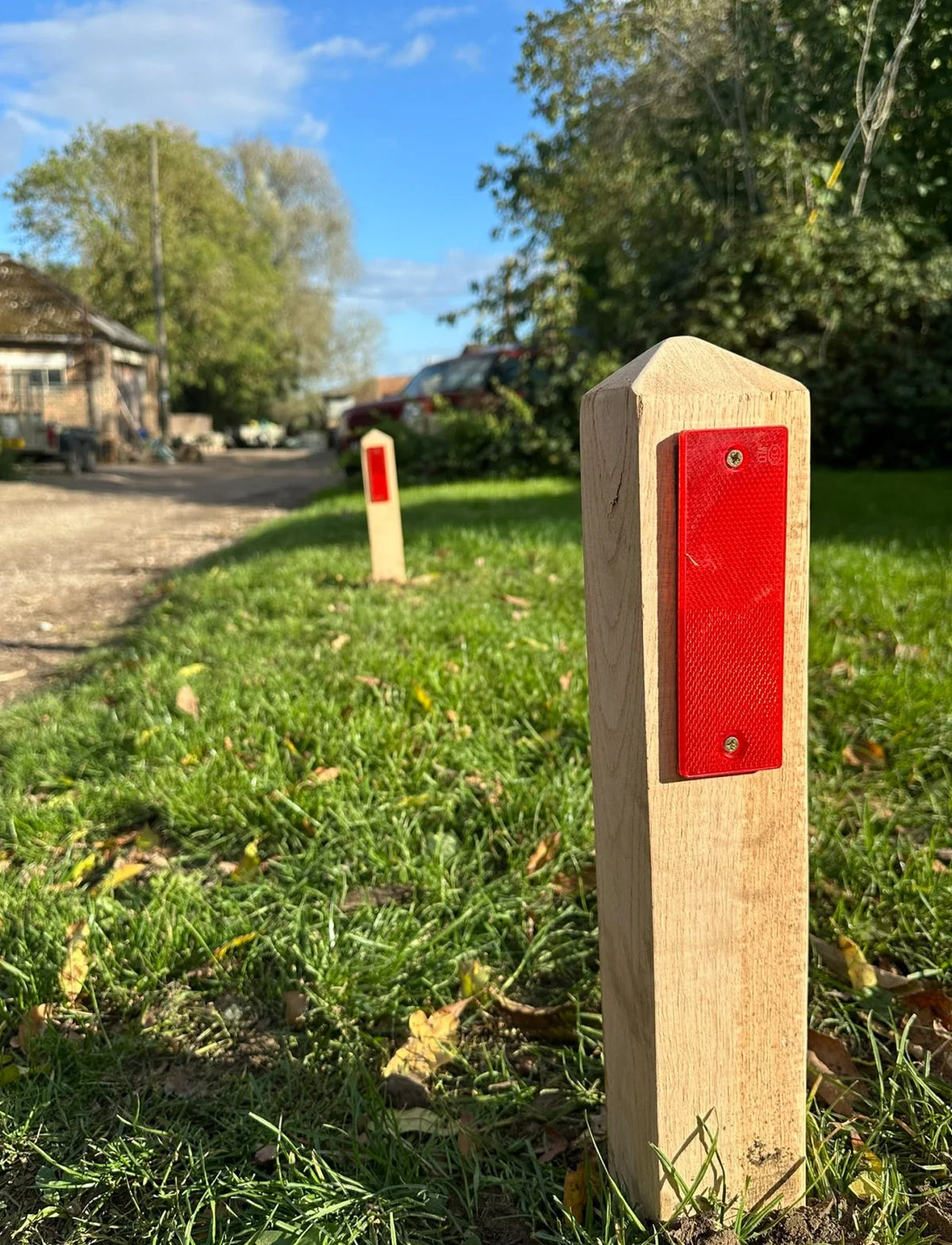 A close-up of a light wood post with a bright red reflective strip in an outdoor grassy area, with a second matching post further in the background near a path and trees.