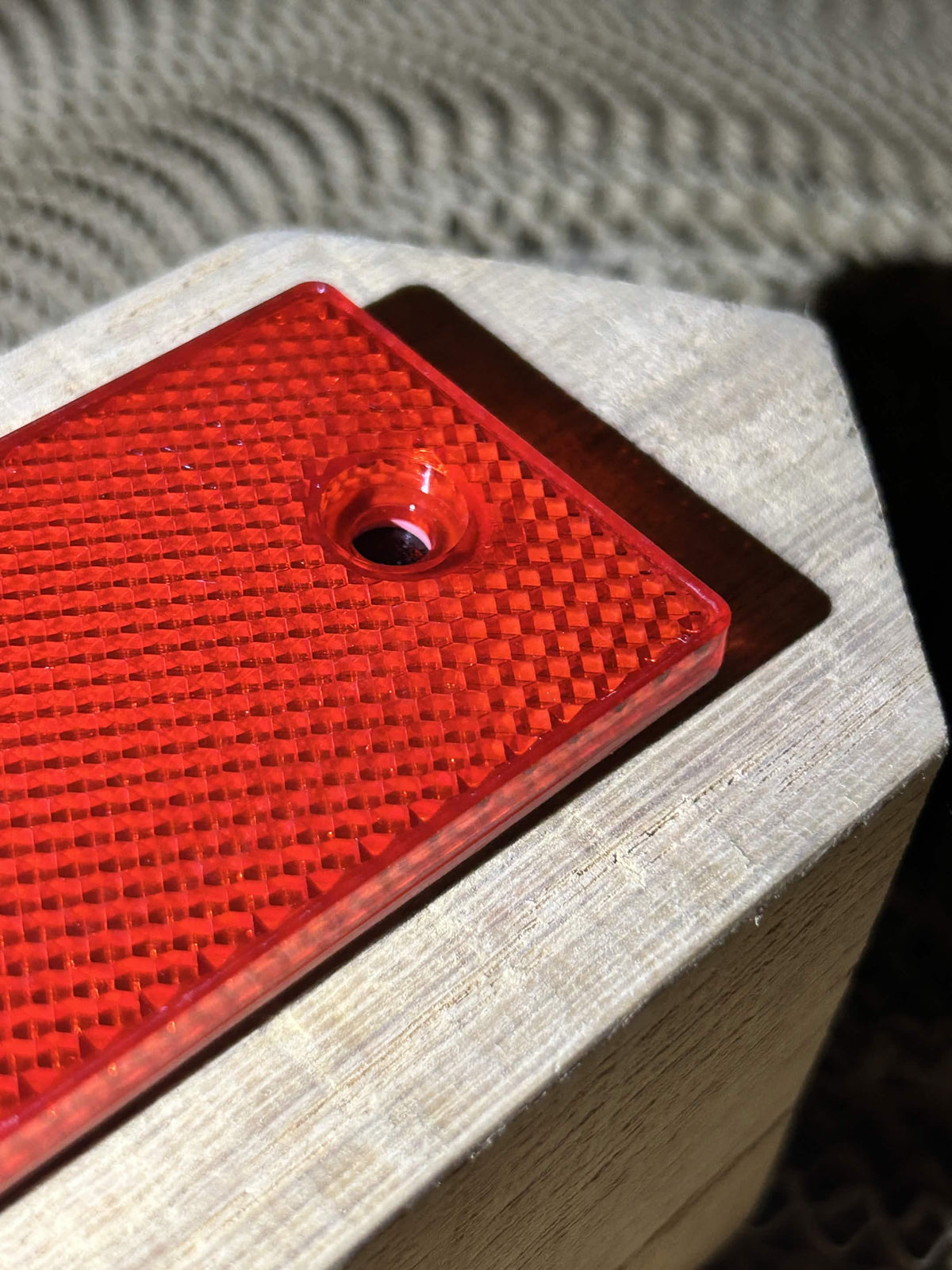 A close-up of a bright red textured square reflector with a mounting hole, mounted on a light-colored wooden block.