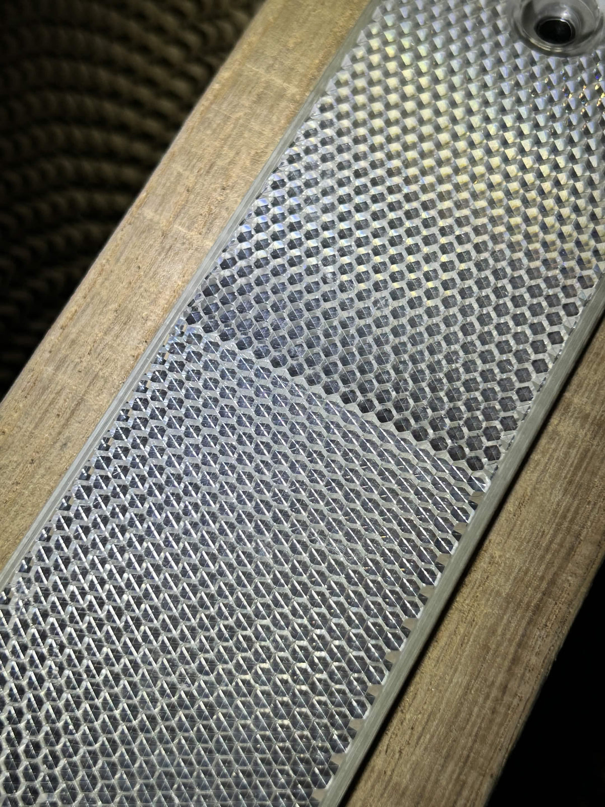 Close-up of a metallic silver mesh or honeycomb-textured panel mounted on a light wood surface, with a dark woven material visible in the upper left.