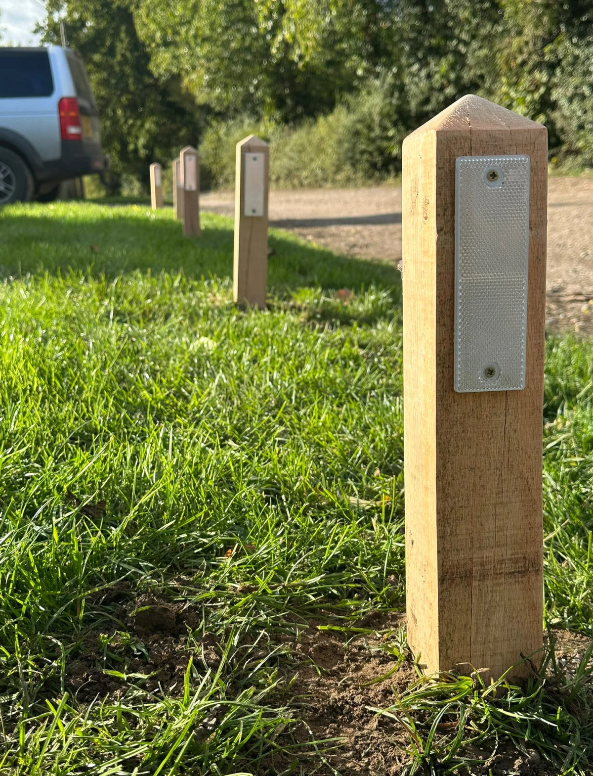 A close-up of a wooden post with a reflective silver panel in a grassy outdoor setting, with similar posts receding into the background near a dirt path and a vehicle.